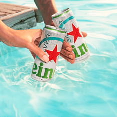 Two cans of Heineken Silver held above bright blue swimming pool water.