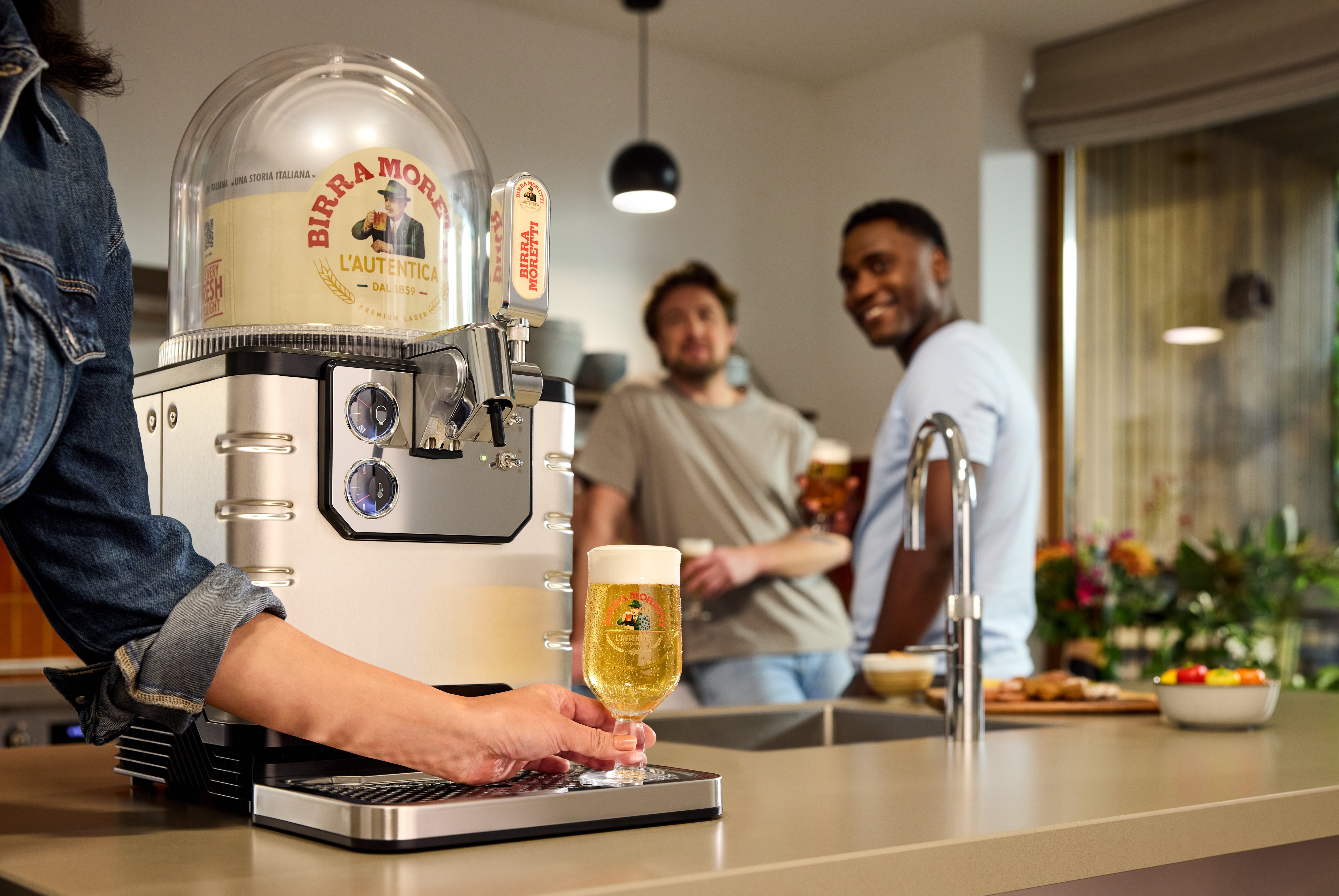 Person using a beer tap to pour a glass of beer in a kitchen setting with two people in the background.