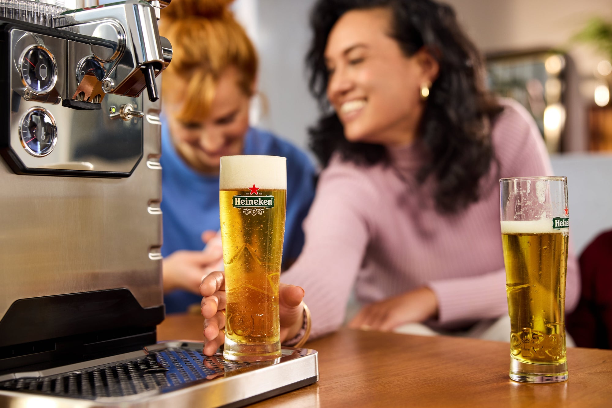Two women enjoying Heineken beer at a bar with a BLADE beer machine in the foreground.