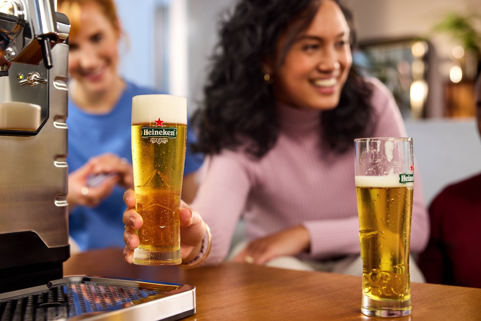 Two glasses of Heineken beer on a bar counter with a blurred background of people and a living room setting.