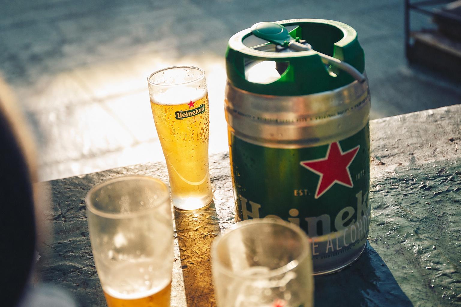 A green and silver Heineken beer keg on a stone table next to two glasses of golden beer, with a blurred outdoor background.