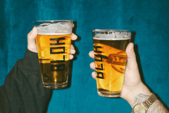 Two hands holding up pint glasses filled with lager against a deep blue background.