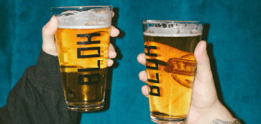 Two hands holding up pint glasses filled with lager against a deep blue background.