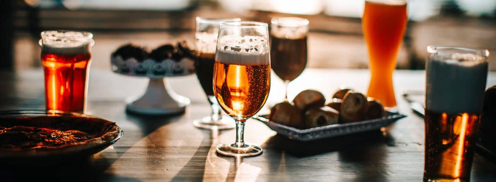 Four glasses of beer on a table with a selection of snacks.