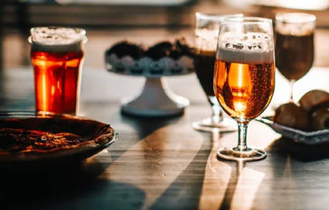 Four glasses of beer on a table with a selection of snacks.