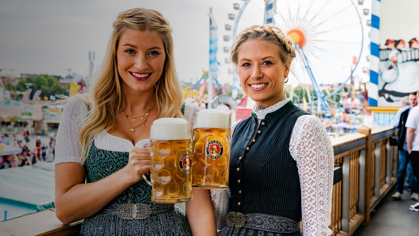 Two women wearing dirndls pose holding steins of Paulaner beer at the Oktoberfest beer festival.