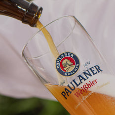 A bottle of beer being poured into a Paulaner Weissbier glass.